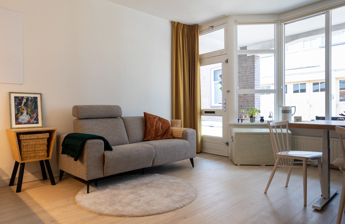 Foto van een grijze bank met daarop een groen kleed en bruin kussentje, daarachter is een witte muur en voor de bank ligt een creme kleurig rond vloerkleed. Je ziet de deur en ramen, met een okergeel gordijn in de hoek hangen.